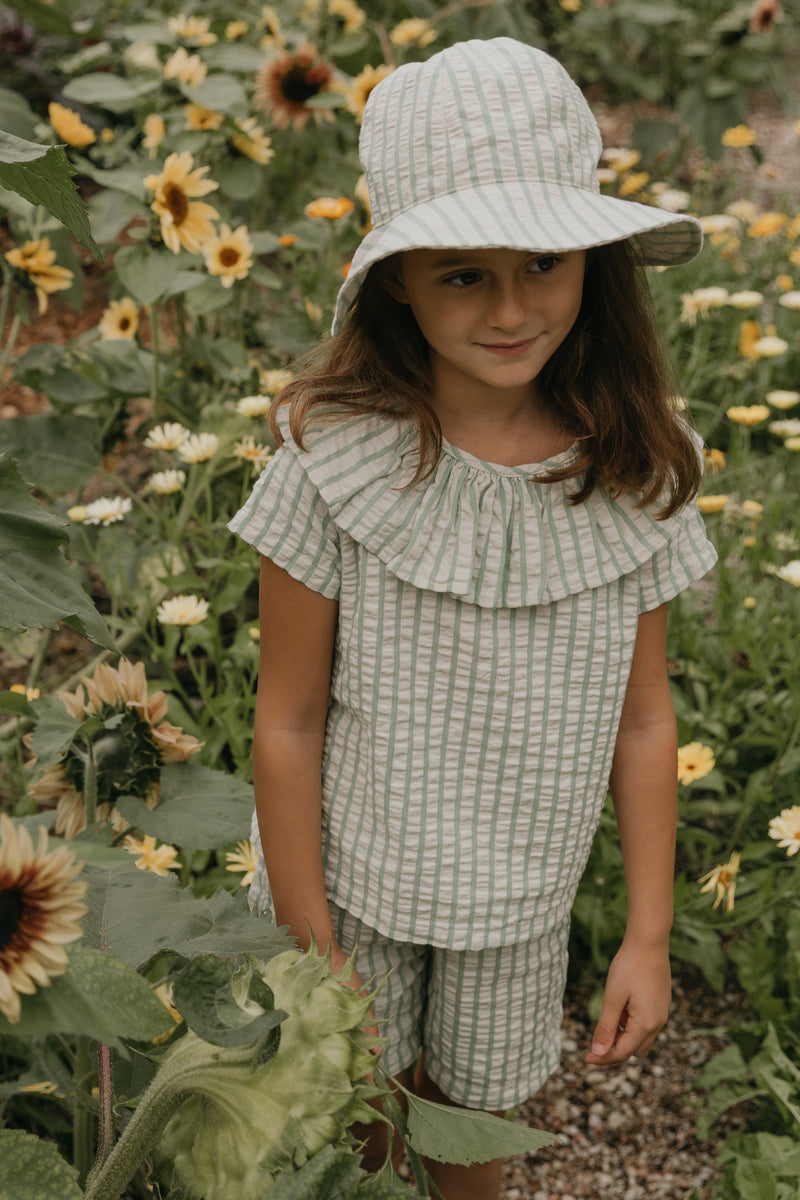 Mädchen gekleidet in Garbo Shorts, beige grün gestreift, seersucker-bio-Baumwolle, stripe emerald muster, Sommerhut. Kind steht in einem Sonnenblumenfeld.