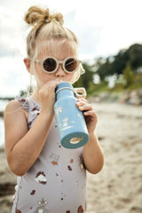 Kleines Mädchen mit heller Sonnenbrille und Badeanzug, trinkt aus einer blauen Trinkflasche mit Maus-Motiven am Strand.