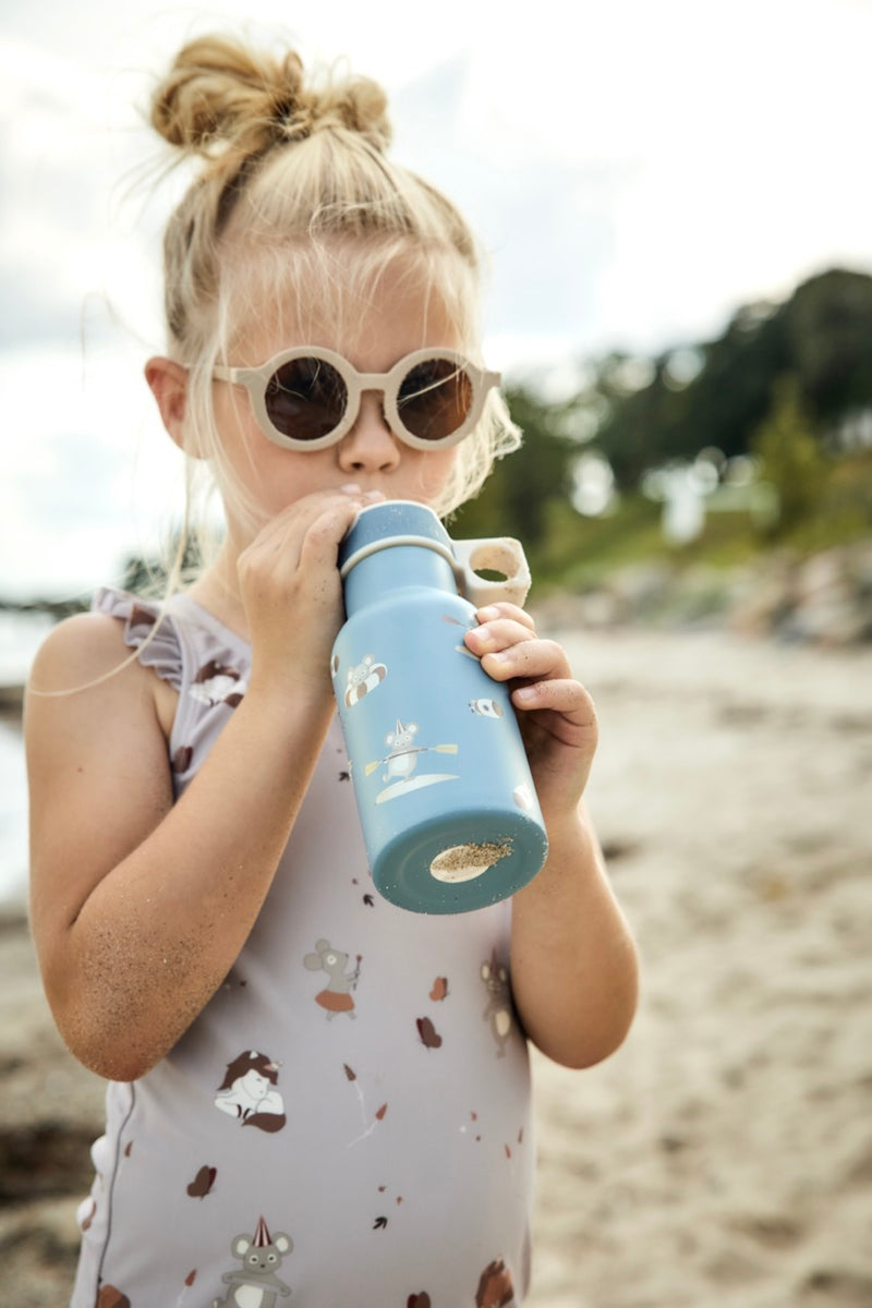 Kleines Mädchen mit heller Sonnenbrille und Badeanzug, trinkt aus einer blauen Trinkflasche mit Maus-Motiven am Strand.