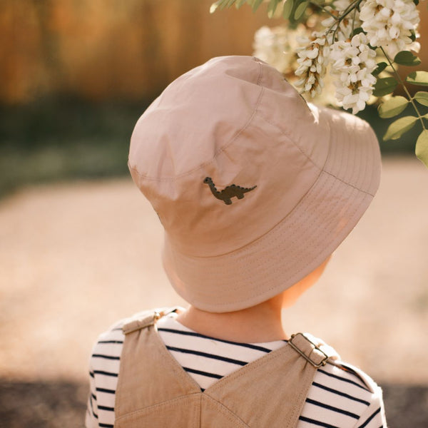 Beiger Baby-Becherhut aus Bio-Baumwolle mit grüner Dino-Stickerei, getragen von einem Kind im gestreiften Shirt und beigefarbener Hose.