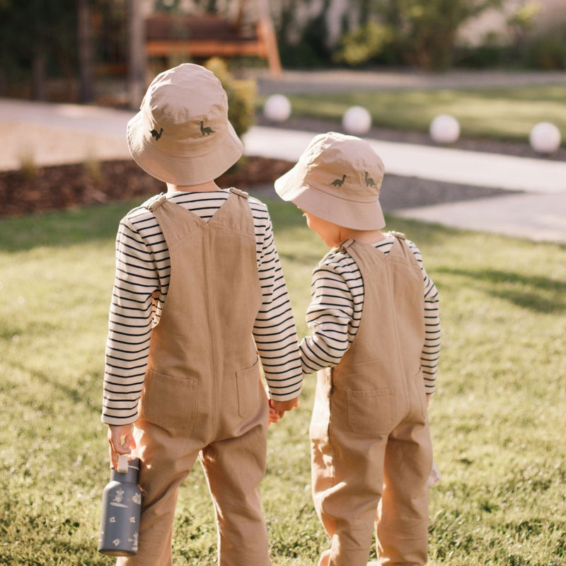 Beiger Baby-Becherhut aus Bio-Baumwolle mit grüner Dino-Stickerei, getragen von einem Kind im gestreiften Shirt und beigefarbener Hose.