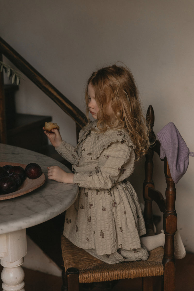 Mädchen trägt ein ähnliches Model vom Langärmeligen Kleid in weicher Baumwoll-Seersucker-Qualität mit schönem handgezeichnetem Druck und Rüsche über den Schultern. Das Kleid hat weiche elastische Bündchen am Ärmelabschluss und Holzimitatknöpfe am Rücken.