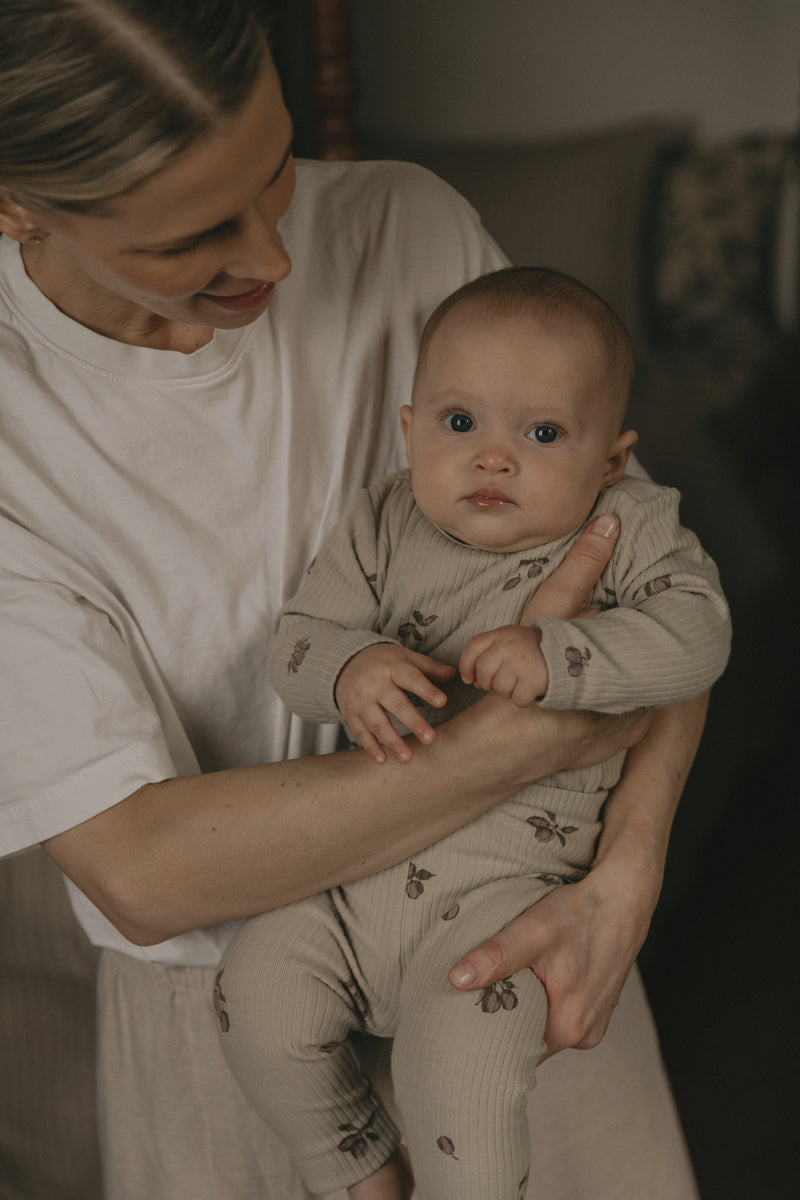 Baby in einem cremefarbenen, floralen Jersey-Onsie von Garbo&Friends, wird von Mama auf dem Schoss gehalten. 
