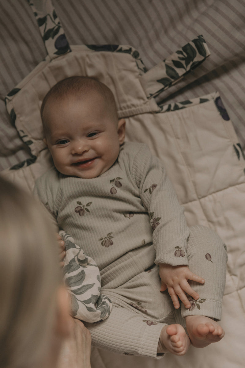 Baby in einem cremefarbenen, floralen Jersey-Onsie von Garbo&Friends, liegend auf einer gemusterten Decke mit Pflanzenmotiven.