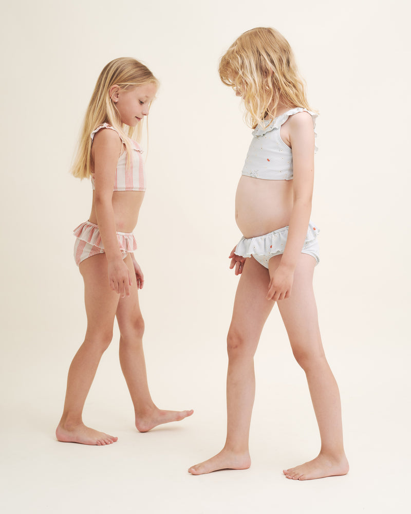 Vollständig gefüttertes Bikini Set mit dekorativen Rüschendetails. Hergestellt aus recycletem, weichen und dehnbaren Material. Kinder am Strand in Bikini in aprico/beige und hellblau.