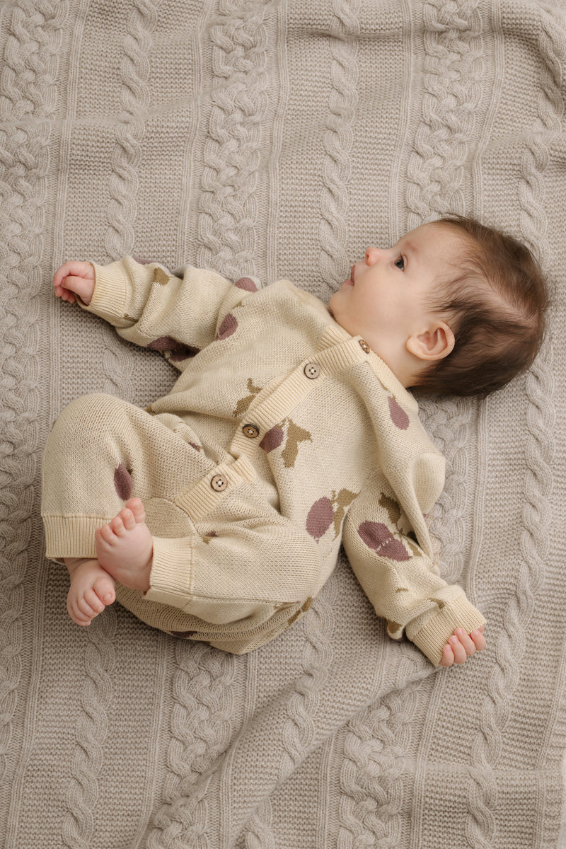 Baby trägt einen gestrickten Onesie. Jacquard Strick-Overall für Babies in weicher Baumwollqualität mit Rundhalsausschnitt, Kokosnussknöpfen und Rippenbündchen am unteren Ende und am Ärmelabschluss.